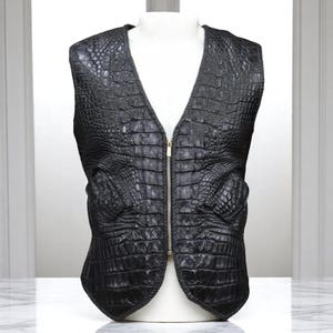 Puede incluir: Un chaleco de cuero negro con textura de piel de cocodrilo. El chaleco tiene cuello en V, cremallera completa y dos bolsillos delanteros. Se exhibe en un maniquí blanco.