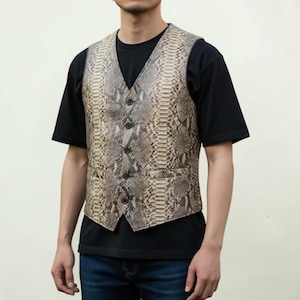 May include: A sleeveless vest featuring a detailed snakeskin pattern in shades of beige, brown, and grey. The garment has a V-neckline, a five-button front closure, and two small welt pockets at the waist, worn over a plain black short-sleeved t-shirt.