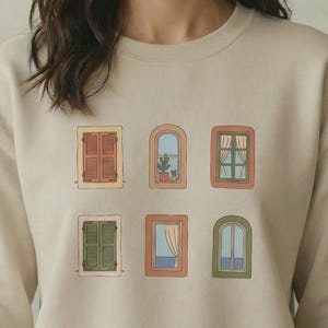 Può includere: Una felpa girocollo color beige con una stampa grafica di sei diverse finestre disposte su due file. Le finestre variano per forma e colore, mostrando diversi stili architettonici, persiane, tende e una piccola pianta in vaso.