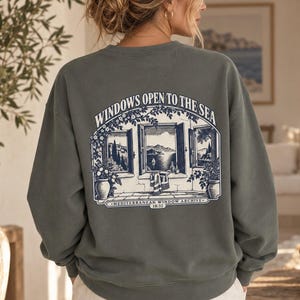 Può includere: Una felpa girocollo verde oliva scuro con una grafica bianca sul retro. Il disegno raffigura una finestra aperta sul mare con il testo "WINDOWS OPEN TO THE SEA" e "MEDITERRANEAN WINDOW ARCHIVE 18:32" stampato in un carattere con grazie.