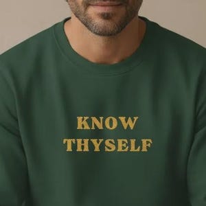 Può includere: Una felpa verde scuro con la scritta dorata "KNOW THYSELF". La felpa girocollo è realizzata in un materiale morbido. È un capo casual, adatto all'uso quotidiano.