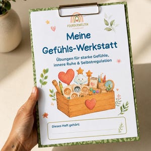 Puede incluir: Un portapapeles con un documento blanco titulado Meine Gefühls-Werkstatt con el subtítulo Übungen für starke Gefühle, innere Ruhe & Selbstregulation. La página presenta una ilustración en acuarela de una caja de madera llena de tarjetas de expresión emocional, un corazón y materiales de arte, con un espacio en la parte inferior etiquetado Dieses Heft gehört.