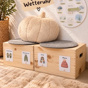 Puede incluir: Dos cajas de almacenamiento de madera clara con cojines de fieltro gris, rematadas con un cojín de felpa beige con forma de calabaza. Un reloj meteorológico circular con texto en alemán y una flecha verde se sitúa encima, junto a + gratis Wetteruhr.