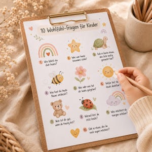 May include: A clipboard with a printed sheet featuring the text "10 Wohlfühl-Fragen für Kinder" and illustrations of a rainbow, star, turtle, bee, flower, sun, bear, ladybug, cloud, and heart. A hand holds a pencil.
