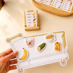 Op de afbeelding: Een spelbord in de vorm van een winkelwagentje met magnetische etenswaren zoals een banaan, yoghurt, komkommer, pasta en sap. Een houten standaard bevat een boodschappenlijstje met de tekst Liste 2, en extra lijstjes liggen op een houten dienblad.
