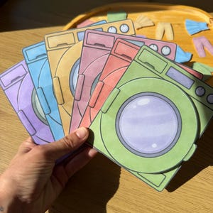 Könnte beinhalten: Ein Set aus Papier-Ausschnitten in Form von Waschmaschinen in verschiedenen Farben wie Grün, Rot, Pink, Gelb, Blau und Lila. Jeder Ausschnitt zeigt eine runde Tür mit einem hellvioletten Zentrum und ein Bedienfeld mit einem Drehknopf oben.