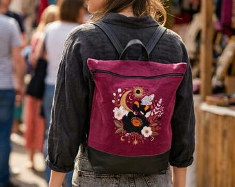 May include: A burgundy canvas backpack with black leather accents and straps. The front features an embroidered design of a bumblebee, a crescent moon, a sun, and various white and black flowers with green leaves and golden star accents.