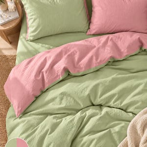 May include: A bedding set featuring a reversible duvet cover and two pillowcases in sage green and soft pink. The fabric has a textured, crinkled appearance. The duvet cover is folded to display both the green and pink sides simultaneously.
