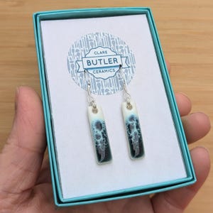 May include: A pair of ceramic earrings in a teal gift box. The earrings are rectangular and have a white and blue glaze with a black pattern. The earrings are hanging from silver hooks. The box has a label that says "Clare Butler Ceramics".