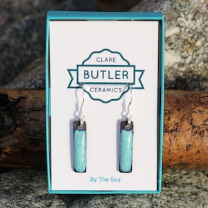 Könnte beinhalten: Ein Paar rechteckige, türkisfarbene und braune Keramikohrringe mit silbernen Haken, präsentiert in einer tealfarbenen Schachtel. Auf der Schachtel steht "Clare Butler Ceramics" und "By The Sea".