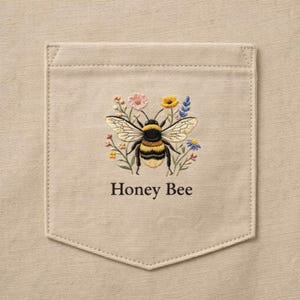 Puede incluir: Un bolsillo beige con un diseño bordado de abeja. La abeja es amarilla y negra, rodeada de flores coloridas. El texto "Honey Bee" está bordado debajo de la abeja.