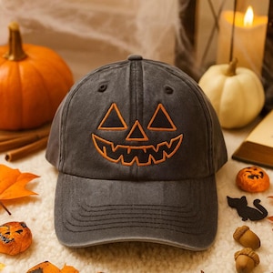 May include: A gray baseball cap with an embroidered jack-o'-lantern face in orange. The cap is surrounded by Halloween decorations, including pumpkins, leaves, and candles, creating a festive autumn scene.