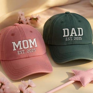 May include: Two baseball caps, one dusty rose and one dark green. The rose cap has "MOM EST. 2025" embroidered in white. The green cap has "DAD EST. 2025" embroidered in white. A pink star wand is in the foreground.