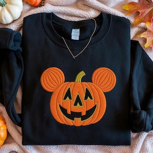 May include: Black sweatshirt featuring an embroidered orange jack-o'-lantern design with mouse ears. The sweatshirt is a crew neck style and is surrounded by pumpkins and fall foliage.