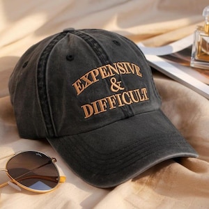 May include: A black baseball cap with the embroidered text "EXPENSIVE & DIFFICULT" in gold. The cap has a curved brim and a distressed look. Sunglasses are visible in the foreground.
