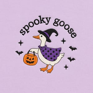 May include: Embroidered design featuring a goose dressed for Halloween. The goose wears a witch hat and cape, carrying a jack-o'-lantern. The words "spooky goose" are arched above the goose, with bats and stars around it. The background is a light purple color.