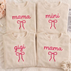 Puede incluir: Cuatro sudaderas color crema con texto bordado en rosa y diseños de lazo. Las sudaderas presentan las palabras "mama", "mini", "gigi" y "mama". Las sudaderas están dispuestas sobre una superficie blanca.