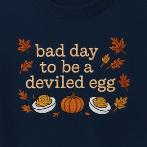 Op de afbeelding: Marineblauwe sweatshirt met de geborduurde tekst "bad day to be a deviled egg". Het ontwerp bevat een pompoen, gevulde eieren en herfstbladeren in oranje en goud.