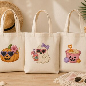 May include: Three off-white canvas tote bags with Halloween-themed designs. One features a pumpkin with heart-shaped sunglasses, another a ghost with heart-shaped sunglasses, and the last a sand pail with a jack-o'-lantern face.