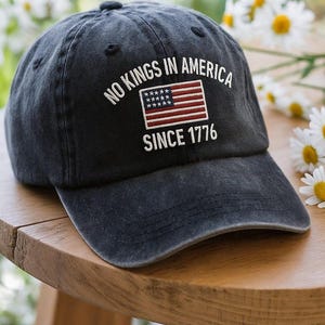 May include: A navy blue baseball cap with the text "NO KINGS IN AMERICA SINCE 1776" and an embroidered American flag. The cap is sitting on a wooden table, with white flowers in the background.