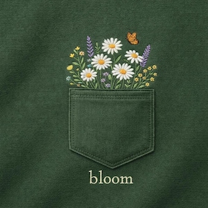 Może przedstawiać: Ciemnozielona bluza z haftowanym wzorem kieszeni. Kieszeń zawiera białe stokrotki, lawendę i małego pomarańczowego motyla. Słowo "bloom" jest wyhaftowane poniżej.