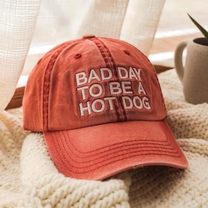 May include: A red baseball cap with the text "BAD DAY TO BE A HOT DOG" embroidered in white. The hat has a curved brim and a distressed look, suggesting a vintage or worn style. The cap is sitting on a cream-colored knitted blanket.