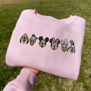 Puede incluir: Una sudadera rosa claro con caras bordadas de personajes de dibujos animados, incluyendo Mickey Mouse, Minnie Mouse, Donald Duck, Daisy Duck, Goofy y Pluto. La sudadera est&aacute; doblada, con los personajes dispuestos en una fila en el pecho.