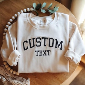 Può includere: Felpa color crema con le parole "CUSTOM TEXT" in blu navy, arcuate sul petto. La parte superiore a maniche lunghe è ripiegata ordinatamente su una superficie di legno, con un rametto di verde e perline decorative sullo sfondo.