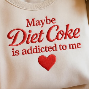 May include: A cream-coloured sweatshirt featuring the embroidered phrase "Maybe Diet Coke is addicted to me" in red script. A red heart is embroidered below the text. The sweatshirt has a crew neck.