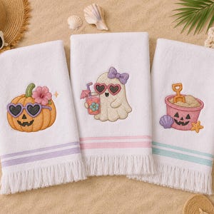 May include: Three white beach towels with embroidered designs. One features a pumpkin with heart-shaped sunglasses, another a ghost with a drink, and the third a sand pail. Each towel has a colored stripe and fringe.