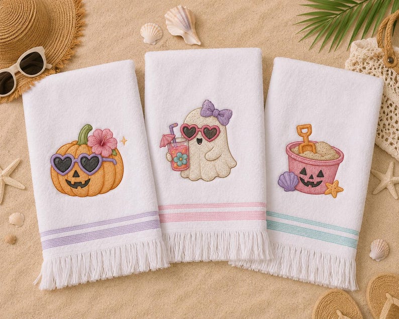 May include: Three white beach towels with embroidered designs. One features a pumpkin with heart-shaped sunglasses, another a ghost with a drink, and the third a sand pail. Each towel has a colored stripe and fringe.