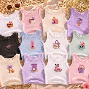 May include: A collection of ribbed tank tops in various colors, each featuring a unique embroidered design. Designs include a pumpkin with sunglasses, a bat with sunglasses, and a flamingo with a witch hat. The text "Summer Spooky Club" is visible.