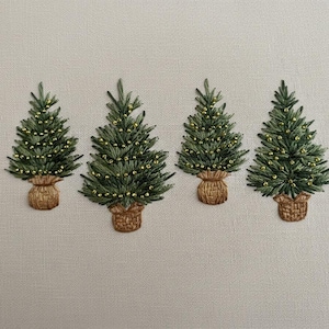 Peut inclure: Quatre sapins de Noël brodés, avec des aiguilles vertes et des lumières dorées, chacun dans un panier en osier. Les arbres sont disposés en rangée sur un fond de tissu beige clair, créant un thème de vacances festif.