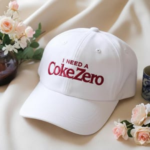 May include: A white baseball cap with the text "I NEED A Coke Zero" embroidered in red. The cap has a curved brim and a rounded crown. The background includes flowers and a decorative cup.