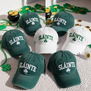 May include: A collection of baseball caps in shades of green and white. Each cap features the word "Sláinte" in white or green lettering, along with a shamrock emblem. The caps are arranged on a textured surface, with decorative gold coins and green beads.