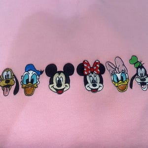 Puede incluir: Tela rosa bordada con seis caras de personajes de dibujos animados: Pluto, Pato Donald, Mickey Mouse, Minnie Mouse, Daisy, y Goofy. Los personajes est&aacute;n en fila, cada uno con colores y rasgos distintivos.