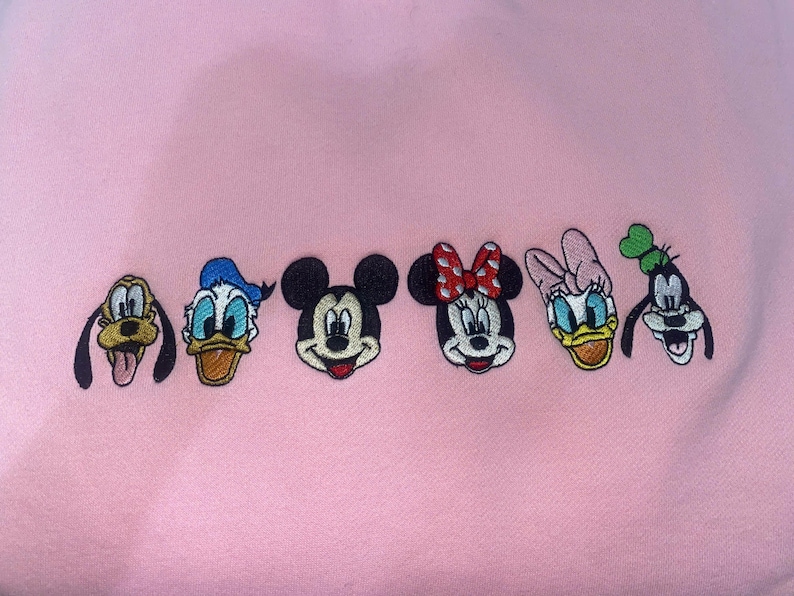 Puede incluir: Tela rosa bordada con seis caras de personajes de dibujos animados: Pluto, Pato Donald, Mickey Mouse, Minnie Mouse, Daisy, y Goofy. Los personajes est&aacute;n en fila, cada uno con colores y rasgos distintivos.