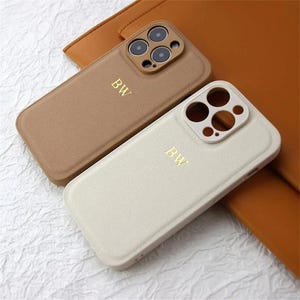 May include: Two leather phone cases, one in tan and the other off-white, are shown. Each case features a gold-coloured "BW" monogram and a camera cutout. The cases are placed on a brown leather surface.