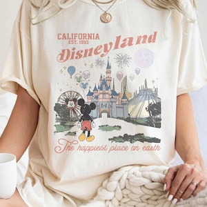 May include: Cream-coloured t-shirt with a Disneyland graphic. The design includes a castle, fireworks, Mickey Mouse, and the text "California Disneyland Est. 1995" and "The happiest place on earth."