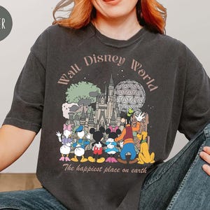 May include: Dark gray t-shirt featuring a Walt Disney World graphic. The design includes the park's logo, iconic characters like Mickey Mouse, and the phrase "The happiest place on earth."