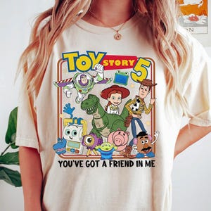 May include: Cream-colored t-shirt featuring a colorful graphic of characters from the animated film "Toy Story 5". The design includes Buzz Lightyear, Woody, Jessie, and other characters, with the text "YOU'VE GOT A FRIEND IN ME".