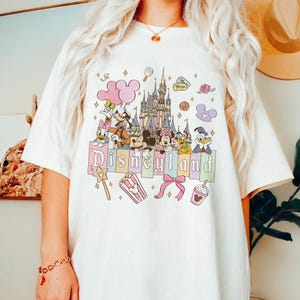 Puede incluir: Camiseta blanquecina con un gr&aacute;fico colorido de Disneyland. El dise&ntilde;o incluye el castillo de Disneyland, Mickey y Minnie Mouse, el Pato Donald y otros personajes de Disney. La palabra "Disneyland" est&aacute; escrita en colores pastel.