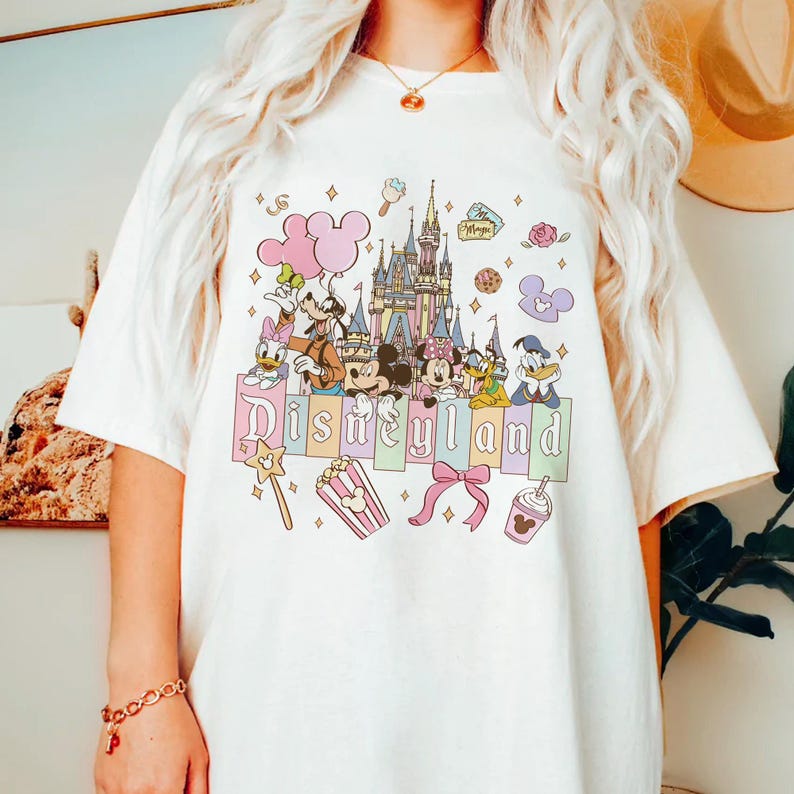 Puede incluir: Camiseta blanquecina con un gr&aacute;fico colorido de Disneyland. El dise&ntilde;o incluye el castillo de Disneyland, Mickey y Minnie Mouse, el Pato Donald y otros personajes de Disney. La palabra "Disneyland" est&aacute; escrita en colores pastel.