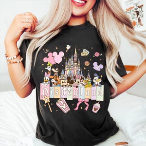 Puede incluir: Camiseta negra con un gr&aacute;fico colorido de Disneyland. El dise&ntilde;o incluye personajes de dibujos animados, un castillo y la palabra "Disneyland" en letras pastel. Gr&aacute;ficos adicionales incluyen una varita, palomitas de ma&iacute;z y un lazo.