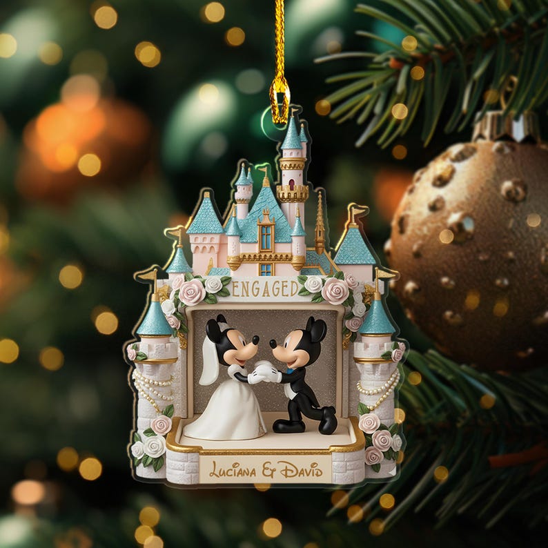 May include: A decorative Christmas ornament featuring Mickey and Minnie Mouse in wedding attire, standing in front of a castle. The ornament is inscribed with the word "ENGAGED" and the names "Luciana & David". The ornament is hanging from a gold string.