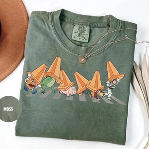 Może przedstawiać: Zielona koszulka Comfort Colors z grafiką postaci z Toy Story przechodzących przez ulicę, z kt&oacute;rych każda ma na sobie pomarańczowy pachołek drogowy. Koszulka ma okrągły dekolt i kr&oacute;tkie rękawy. Złoty naszyjnik jest zarzucony na koszulkę.