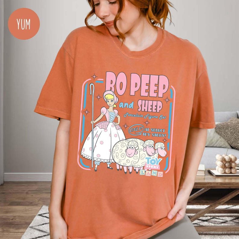 以下が含まれることがあります： オレンジ色のTシャツ。トイ・ストーリーのボー・ピープと羊の漫画のグラフィックが特徴です。「Bo Peep and Sheep」と「Toy Story Land」の文字が含まれています。イラストはパステルカラーの漫画スタイルです。
