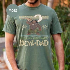 May include: Moss green t-shirt featuring a cartoon illustration of a Polynesian character holding a hook. The shirt has the text "JUST AN ORDINARY" and "DEMI-DAD" in a gold color.