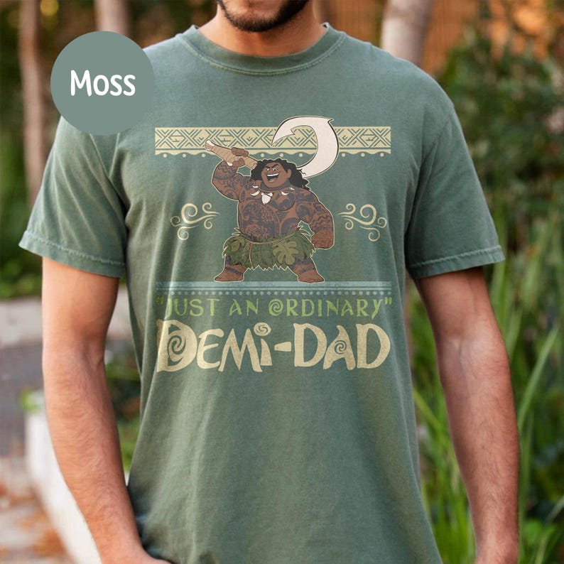 May include: Moss green t-shirt featuring a cartoon illustration of a Polynesian character holding a hook. The shirt has the text "JUST AN ORDINARY" and "DEMI-DAD" in a gold color.