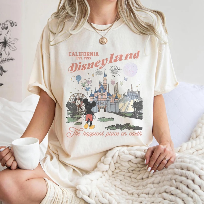 May include: Cream-coloured t-shirt with a Disneyland graphic. The design includes a castle, fireworks, Mickey Mouse, and the text "California Disneyland Est. 1995" and "The happiest place on earth."